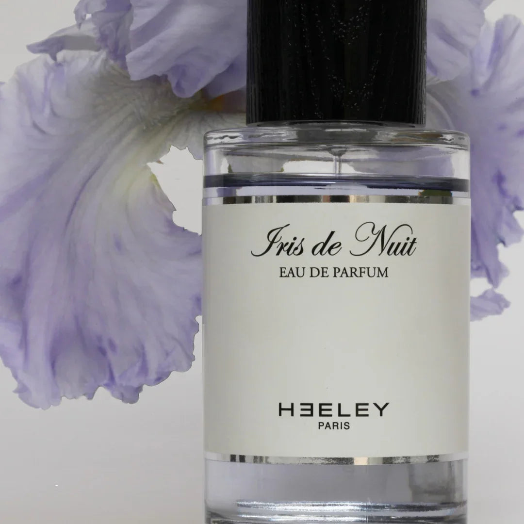 Heeley Iris de Nuit Eau de Parfum in einer durchsichtigen Flasche mit schwarzem Verschluss, Blumenmotiv im Hintergrund.