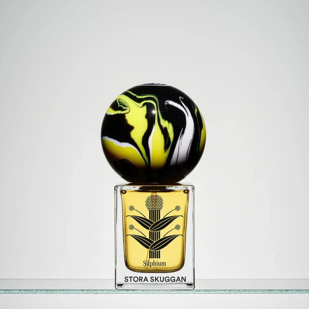 Duftflakon mit kugelförmigem Deckel in Schwarz und Gelb. Klare Glasflasche mit gelbem Inhalt, "Silphium" und "Stora Skuggan" aufgedruckt.