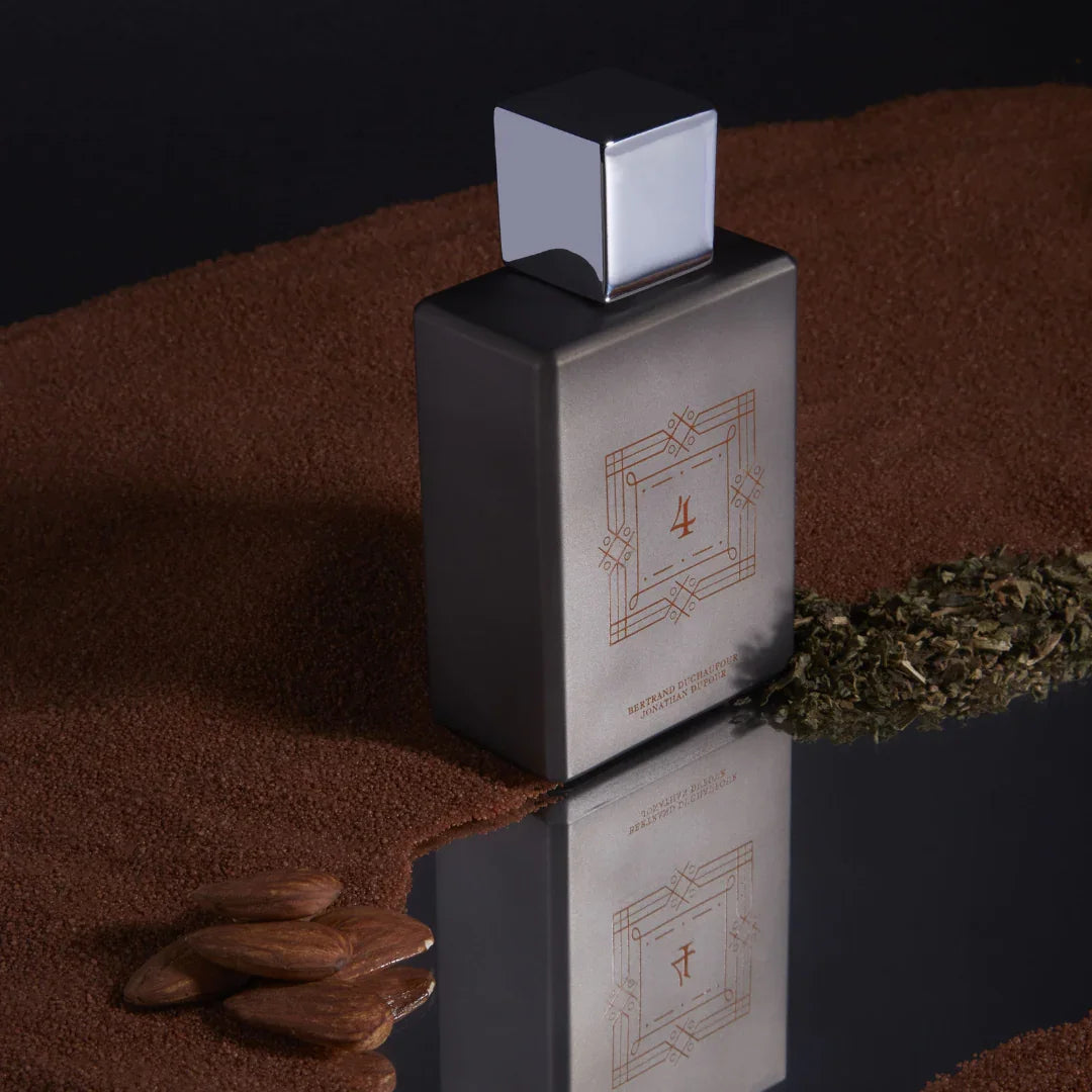 Spiritum Paris BUILDER OF FUTURE Eau de Parfum – Flakon von schräg oben, auf einer spiegelnden Fläche, eingerahmt von Mandeln und braunem Sand – eine sinnliche Einladung, die Grenzen zwischen Natur und Vision bei scent amor zu erkunden