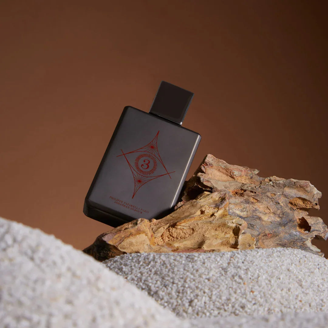 Spiritum Paris WISDOM UNIVERSE Eau de Parfum – Flakon ruht geneigt an einem Stück Holz im Sand, eine meditative Szene, die die Verbindung von Natur, Zeit und Duftkunst spürbar macht – entdecke es bei scent amor