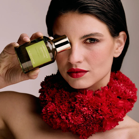 Laboratorio Olfattivo IN_CARNATION Eau de Parfum, Frau mit roten Lippen und floraler roten Halskrause hält den Duftflakon vor ihr Gesicht, erhältlich bei scent amor.