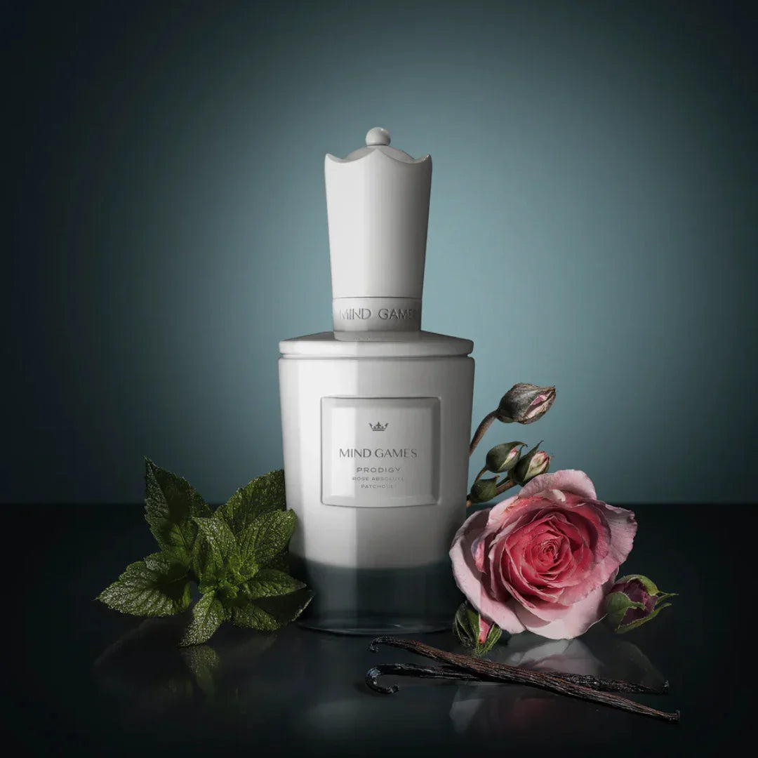 Weißer Flakon des Prodigy Eau de Parfum von Mind Games, dekoriert mit einer Rose, Vanilleschoten und Minzblättern.