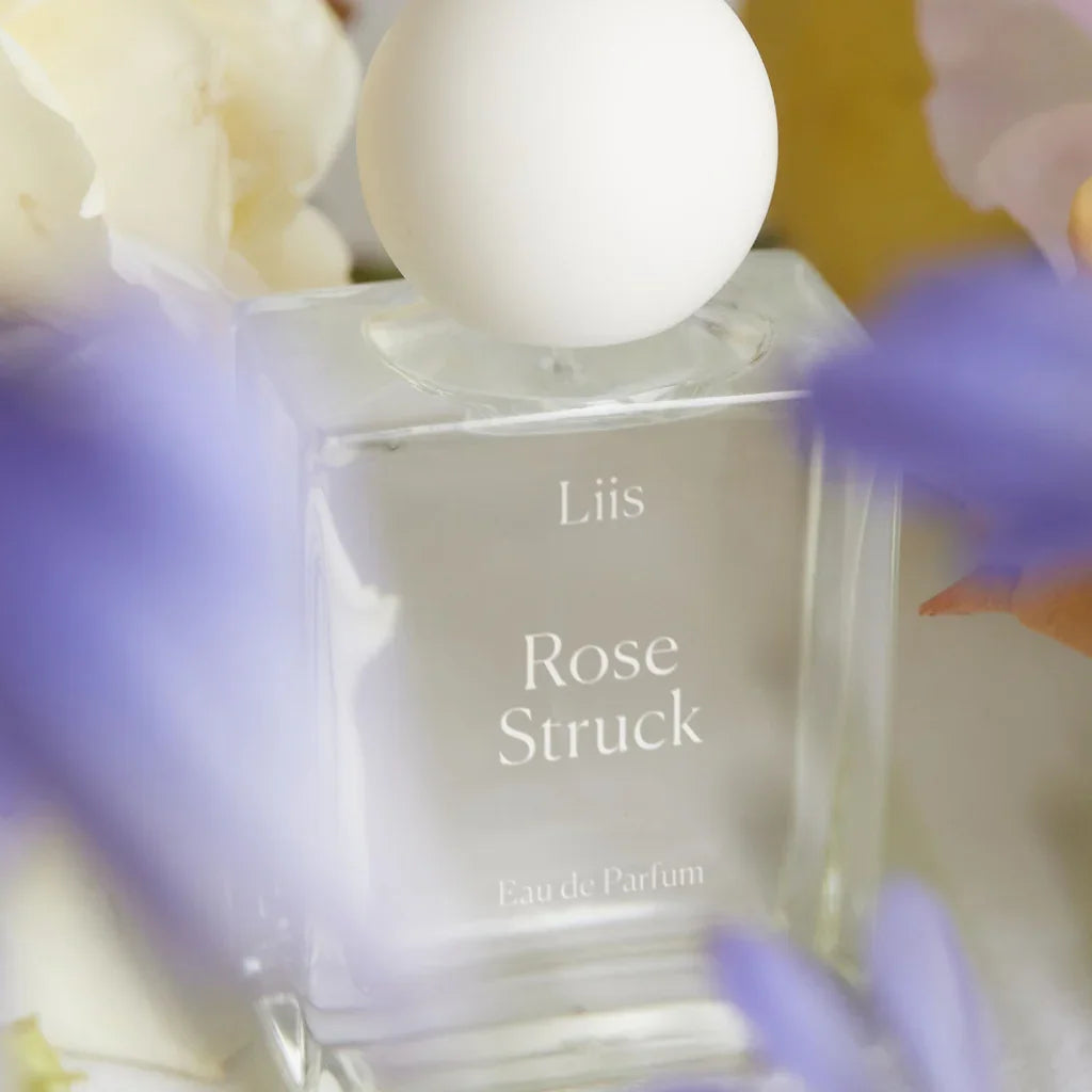 Klares, quadratisches Parfümflakon mit weißer Kugel als Verschluss, "Liis Rose Struck" aufgedruckt. Umgeben von Blumen.