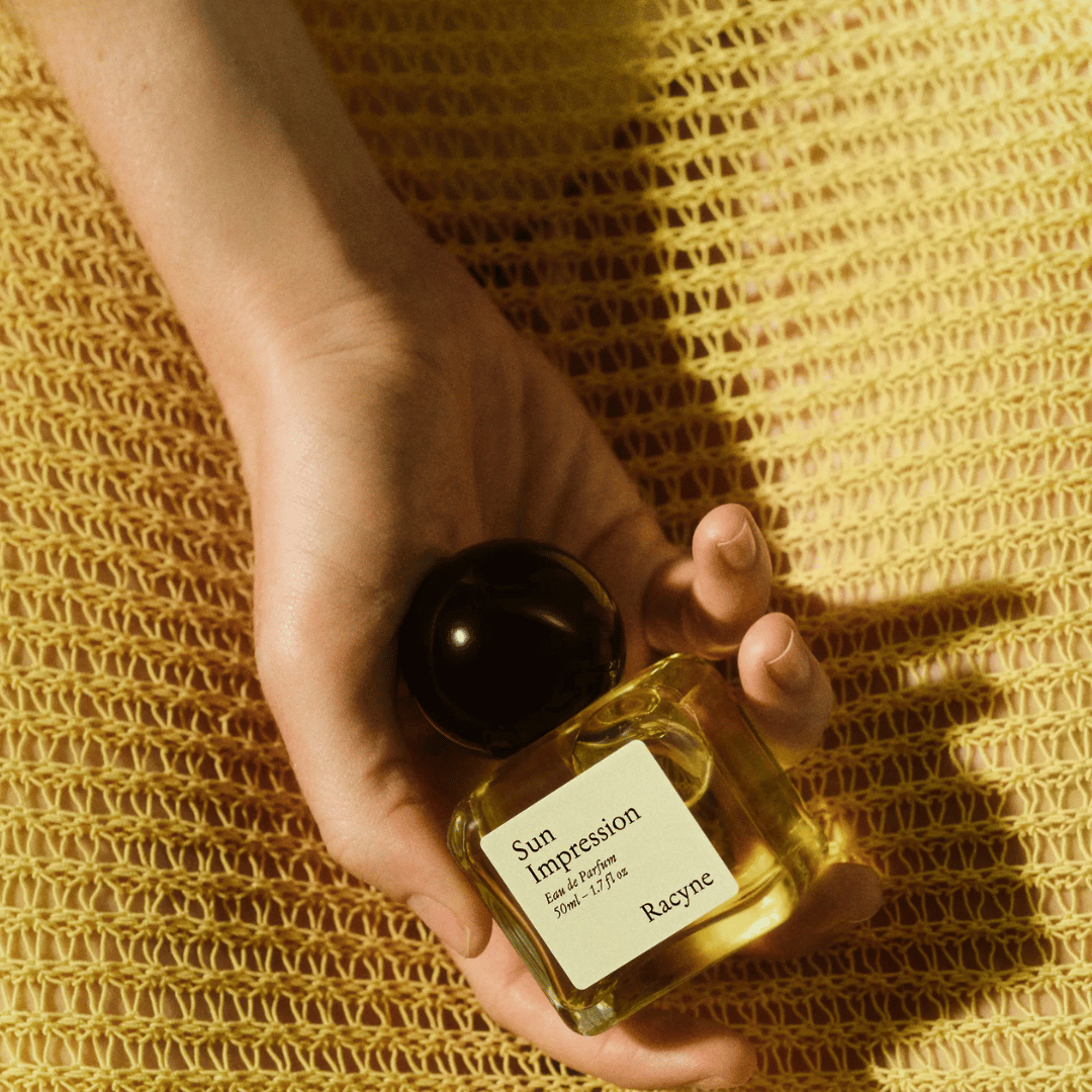 Racyne SUN IMPRESSION Eau de Parfum in Frauenhand vor gelbem Stoff  bei scent amor Fachmann für feine Nischenparfums
