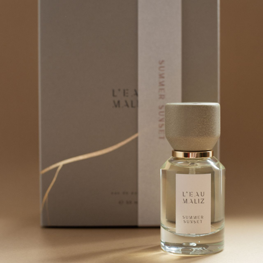 L’Eau Maliz Summer Sunset Eau de Parfum, Flakon vor Originalverpackung in warmer Studio-Szene, zeitloses Design, erhältlich bei scent amor.