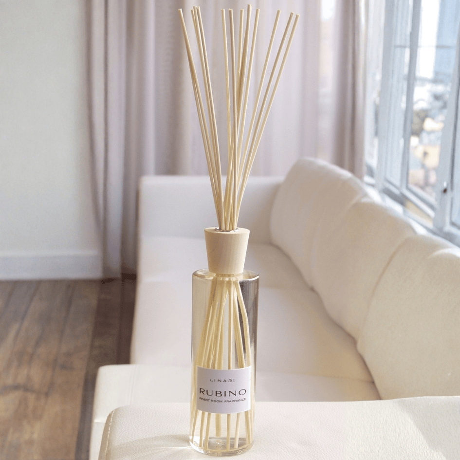 Linari Rubino Diffuser, stilvoller Raumduft-Flakon mit Duftstäbchen auf einem Sofa im hellen Wohnraum, warm und einladend, erhältlich bei scent amor.